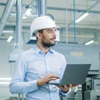 Chief Engineer in the Hard Hat Walks Through Light Modern Factory While Holding Laptop. Successful, Handsome Man in Modern Industrial Environment.