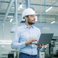 Chief Engineer in the Hard Hat Walks Through Light Modern Factory While Holding Laptop. Successful, Handsome Man in Modern Industrial Environment.