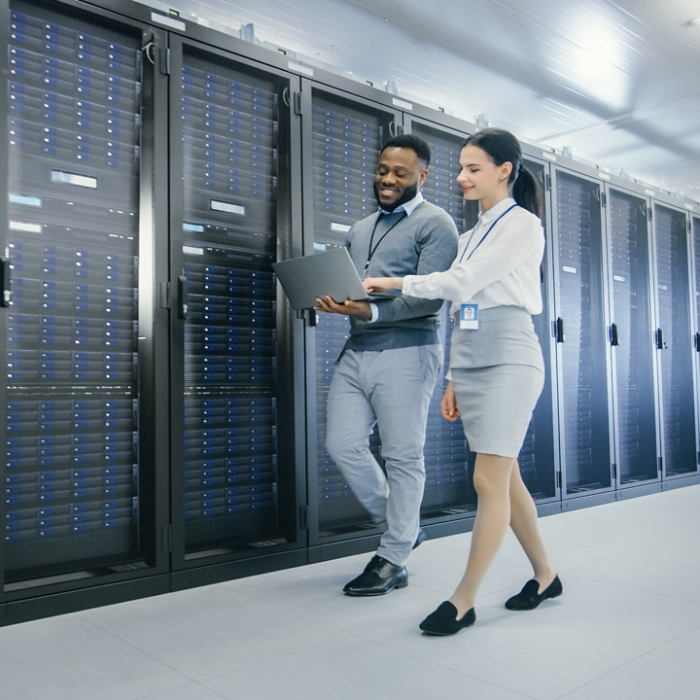 Black IT Technician with a Laptop Computer Gives a Tour to a Young Intern. They Talk in Data Center while Walking Next to Server Racks. Running Diagnostics or Doing Maintenance Work. 