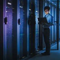 In Dark Data Center: Male IT Specialist Stands Beside the Row of Operational Server Racks, Uses Laptop for Maintenance. Concept for Cloud Computing, Artificial Intelligence, Supercomputer, Cybersecurity. Neon Lights
