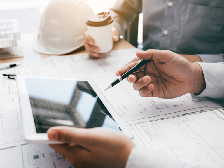 Two engineering working together and using digital tablet looking blueprint and analysis with architectural plan on desk.