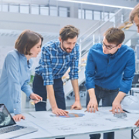 In the Industrial Engineering Facility: Diverse Group of Engineers and Technicians on a Meeting Gather Around Table with Engine Design Technical Drafts, Have Discussion, Analyse Technology