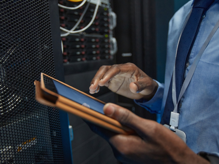 Close up of African American man holding digital tablet while standing by server cabinet and working with supercomputer in data center, copy space