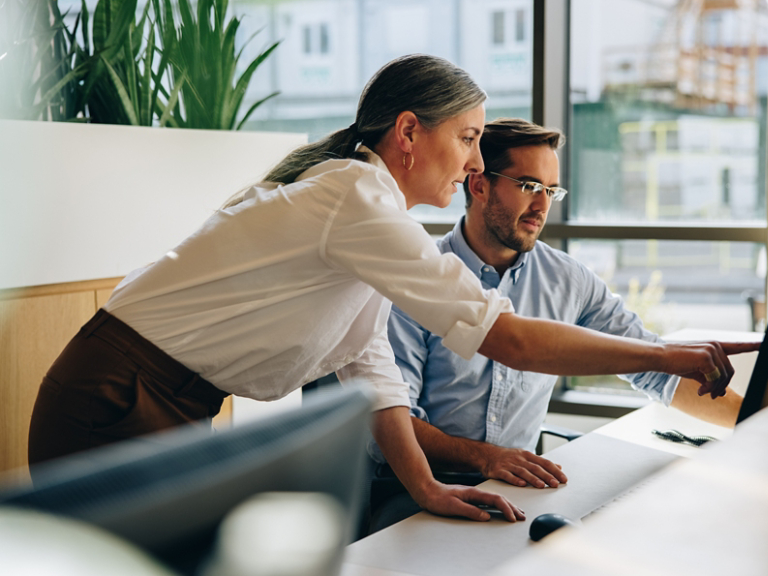 Mature woman working with young man sitting at desk and showing something on computer screen in office. Female executive pointing at desktop monitor and talking with male colleague in office.