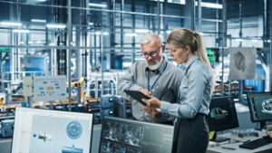 Car Factory Office: Manager Talks with Senior Engineer, Use Tablet Computer to Design Production Conveyor for Advanced Power Engines. Automated Robot Arm Assembly Line Manufacturing Vehicles