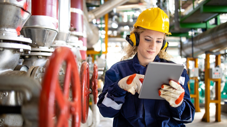 Refinery worker with hardhat and earmuffs checking parameters on tablet computer for oil and gas production.