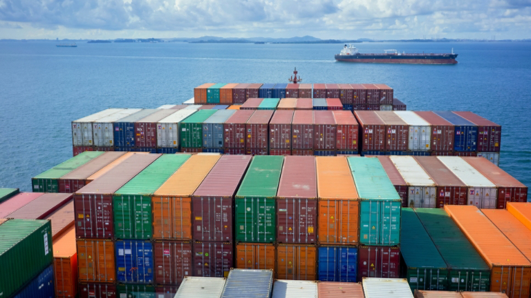 Aerial view of colorful shipping containers stacked on a cargo ship with an expansive ocean and a cloudy sky in the background.