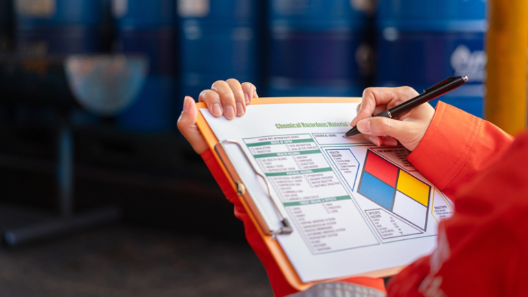 A safety officer is checking on the hazardous material checklist form with chemical storage area at the factory as background. Industrial safety working scene, selective focus.