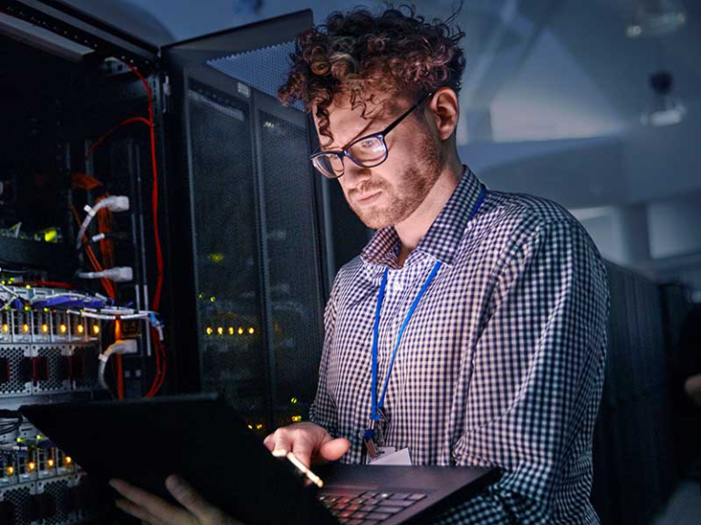 Focused male IT technician working at laptop in dark server room