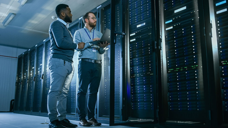 Bearded IT Technician in Glasses with a Laptop Computer and Black Male Engineer Colleague are Talking in Data Center while Working Next to Server Racks. Running Diagnostics or Doing Maintenance Work. 