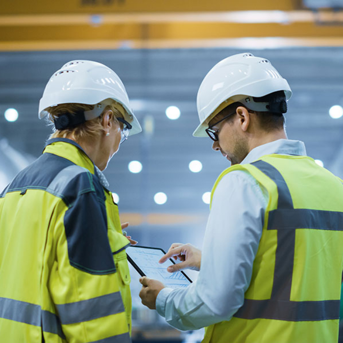 Two Heavy Industry Engineers Stand in Pipe Manufacturing Factory, Use Digital Tablet Computer, Have Discussion. Large Pipe Assembled. Design and Construction of Oil, Gas and Fuels Transport Pipeline