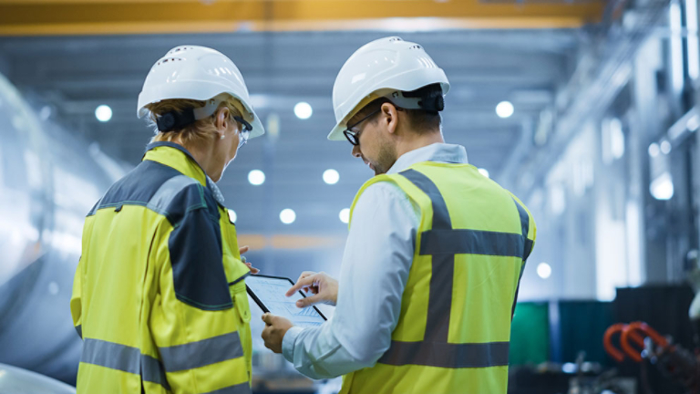 Two Heavy Industry Engineers Stand in Pipe Manufacturing Factory, Use Digital Tablet Computer, Have Discussion. Large Pipe Assembled. Design and Construction of Oil, Gas and Fuels Transport Pipeline