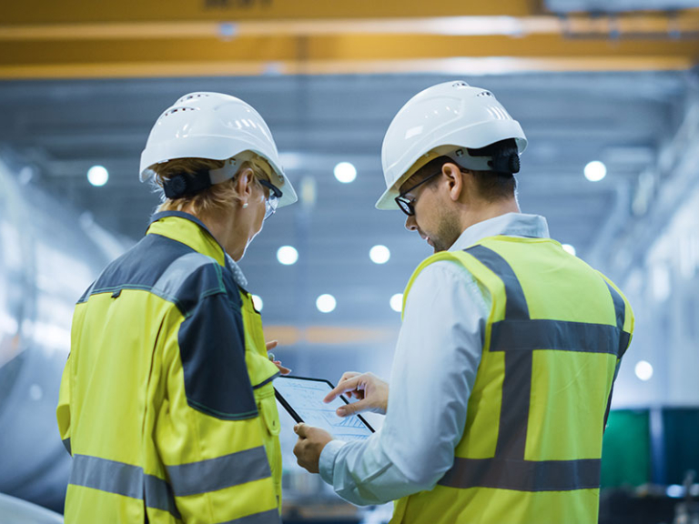 Two Heavy Industry Engineers Stand in Pipe Manufacturing Factory, Use Digital Tablet Computer, Have Discussion. Large Pipe Assembled. Design and Construction of Oil, Gas and Fuels Transport Pipeline