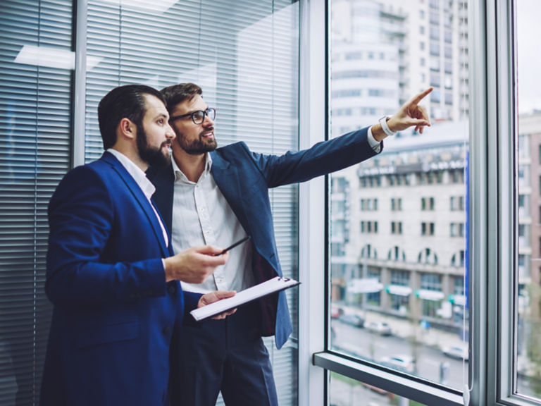 Two caucasian business partners looking in window for building and thinking about making investment, successful entrepreneur pointing outdoors to male colleague standing near with paper folder in hand