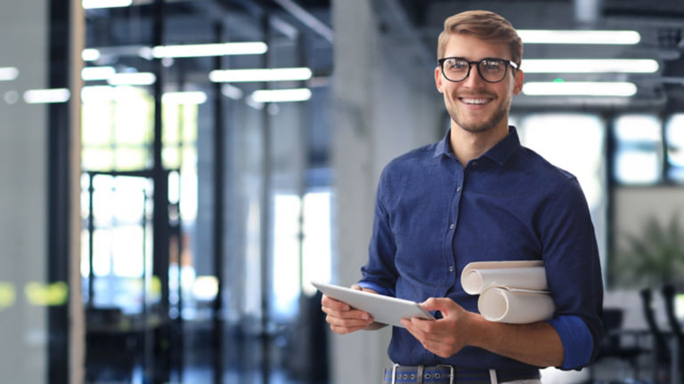 Confident young business man in shirt examining blueprint while standing against a window at office.