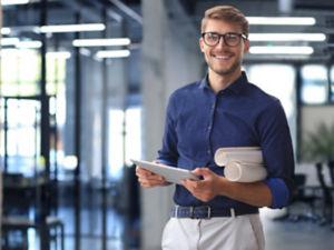 Confident young business man in shirt examining blueprint while standing against a window at office.