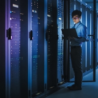 In Dark Data Center: Male IT Specialist Stands Beside the Row of Operational Server Racks, Uses Laptop for Maintenance. Concept for Cloud Computing, Artificial Intelligence, Supercomputer, Cybersecurity. Neon Lights