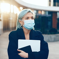 Careful caucasian businesswoman holding a laptop and tablet while wearing a protective mask