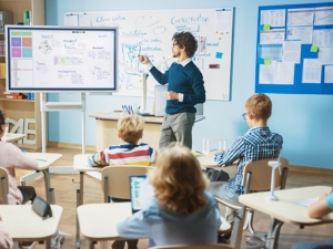 Elementary School Science Teacher Uses Interactive Digital Whiteboard to Show Classroom Full of Children how Software Programming works for Robotics. Science Class, Curious Kids Listening Attentively