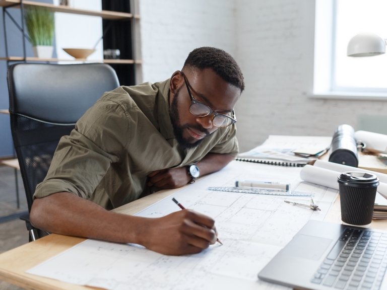Afro-American architect working in office with blueprints.Engineer inspect architectural plan, sketching a construction project. Portrait of black handsome man sitting at workplace. Business concept