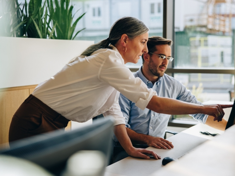 Mature woman working with young man sitting at desk and showing something on computer screen in office. Female executive pointing at desktop monitor and talking with male colleague in office.