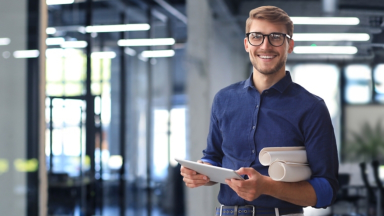 Confident young business man in shirt examining blueprint while standing against a window at office.