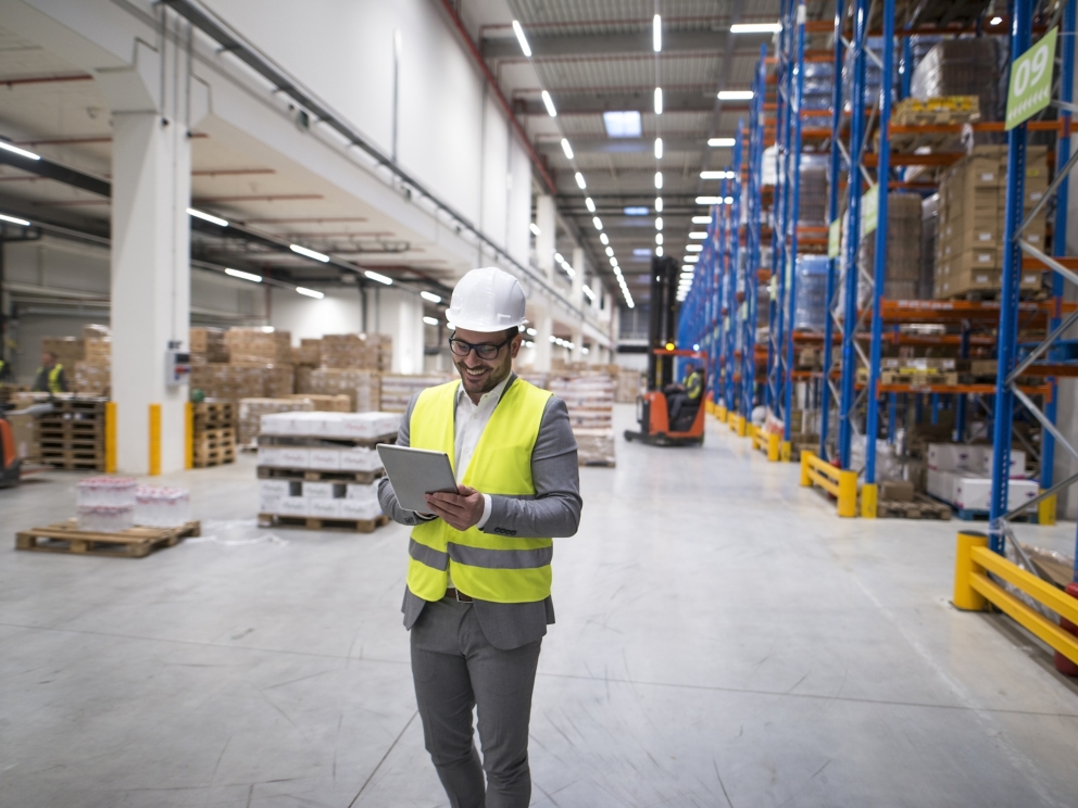 Warehouse manager walking through large storage area and holding tablet while forklift operating in background.