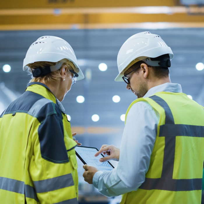 Two Heavy Industry Engineers Stand in Pipe Manufacturing Factory, Use Digital Tablet Computer, Have Discussion. Large Pipe Assembled. Design and Construction of Oil, Gas and Fuels Transport Pipeline
